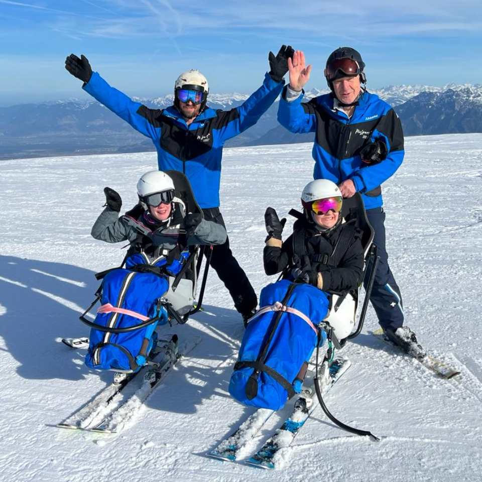 Zwei Personen sitzen lächelnd in adaptiven Skistühlen, während zwei hinter ihnen stehende Guides auf einem schneebedeckten Berghang mit klarem Himmel und Bergen im Hintergrund ihre Arme zum Jubeln heben.