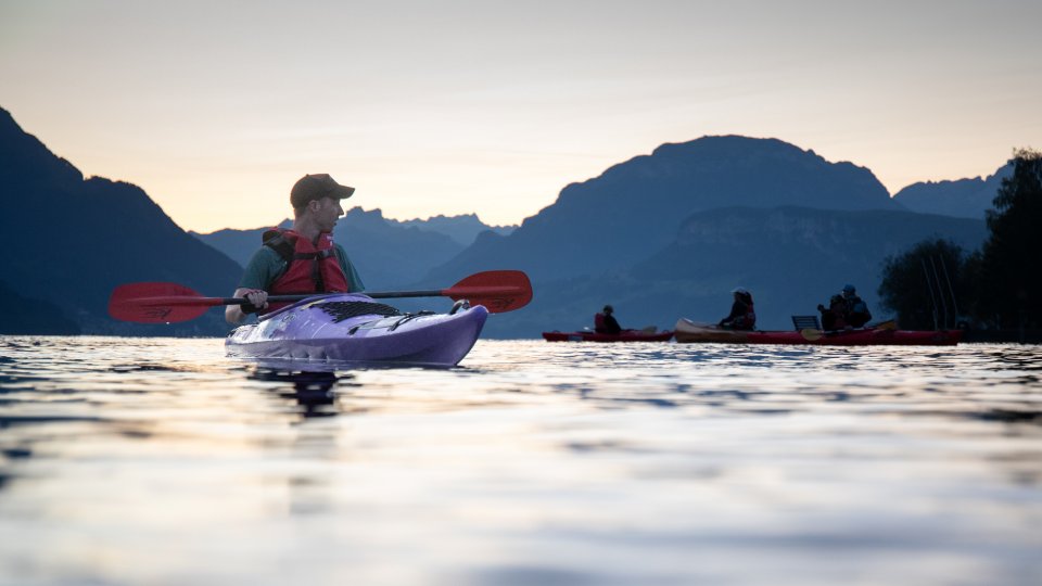 Eine Person in einem violetten Kajak paddelt bei Sonnenuntergang auf einem ruhigen See, im Hintergrund sind Berge zu sehen, und in der Ferne sind zwei weitere Kajakfahrer zu erkennen.