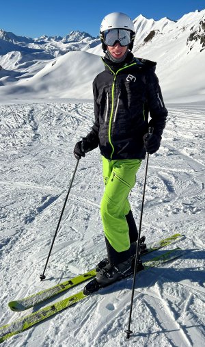 Ein Skifahrer in einer schwarzen Jacke, hellgrünen Hosen und einem Helm steht mit Skistöcken auf verschneiten Hängen, umgeben von Bergen unter einem strahlend blauen Himmel.
