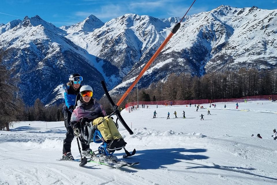 Eine Person in einer angepassten Skiausrüstung wird von einem Skilehrer auf einem verschneiten Berghang unterstützt, im Hintergrund sind Skifahrer und schneebedeckte Gipfel vor einem strahlend blauen Himmel zu sehen.