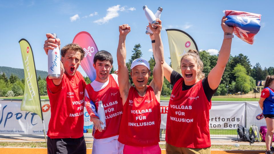 Vier lächelnde Sportler in roten Trikots stehen gemeinsam im Freien und recken jubelnd die Arme in die Höhe. Sie halten Jonglierkeulen und eine Fahne in den Händen, im Hintergrund sind bunte Banner und ein sonniger Himmel zu sehen.