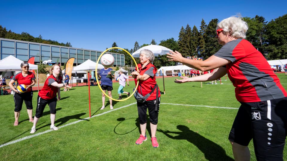 Drei Frauen in roten T-Shirts spielen im Freien und werfen einen Ball durch einen gelben Reifen. Im Hintergrund sind an einem sonnigen Tag weitere Menschen und Zelte zu sehen.