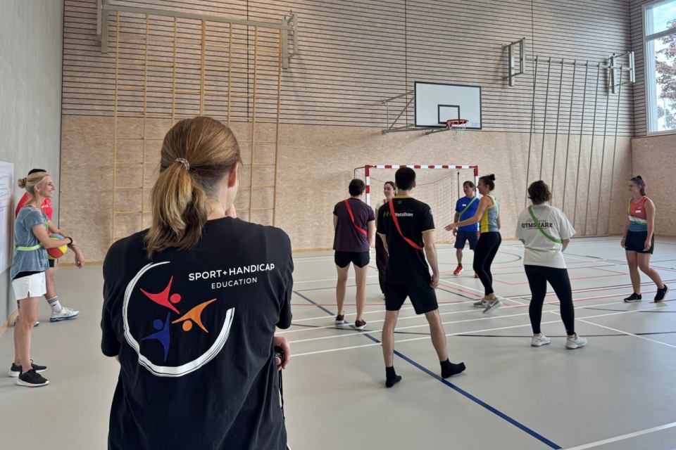 Eine Gruppe von Menschen in Sportkleidung steht in einer Sporthalle und hört sich Anweisungen an. Eine Person im Vordergrund trägt ein schwarzes T-Shirt mit dem Logo von „Sport Handicap Education“ auf dem Rücken.