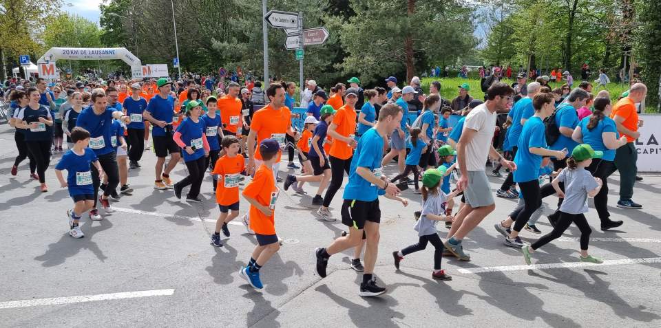 Eine große Gruppe von Menschen, darunter Erwachsene und Kinder in orangefarbenen und blauen Hemden, läuft an einem sonnigen Tag gemeinsam bei einem Straßenrennen, umgeben von Bäumen und Zuschauern.