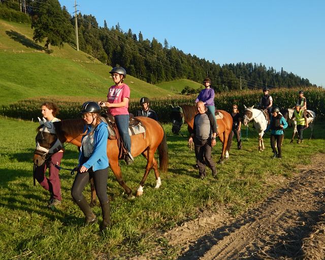 Eine Gruppe von Menschen reitet auf Pferden einen grasbewachsenen Weg entlang, begleitet von anderen, die neben ihnen gehen. Die Szene spielt sich in einer grünen, hügeligen Landschaft unter blauem Himmel ab.