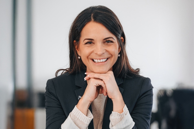Eine Frau mit langen dunklen Haaren lächelt warm und stützt ihr Kinn auf ihre Hände. Sie trägt einen dunklen Blazer und eine helle Bluse und sitzt in einer hellen, modernen Inneneinrichtung.