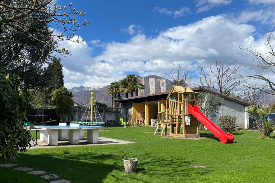 Sonniger Hinterhof mit grünem Gras, einem Holzspielgerät mit einer roten Rutsche, einer Tischtennisplatte und einem Klettergerüst. Im Hintergrund sind Bäume und Berge unter einem teilweise bewölkten Himmel zu sehen.