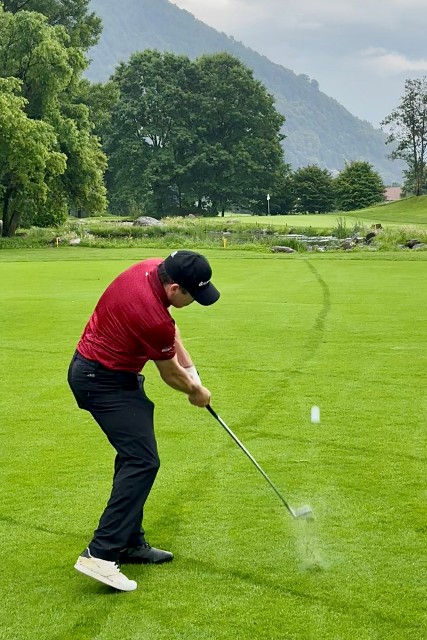 Ein Golfer in rotem Hemd und schwarzer Hose schwingt einen Schläger und schlägt einen Golfball auf einem üppig grünen Fairway, mit Bäumen und Bergen im Hintergrund.