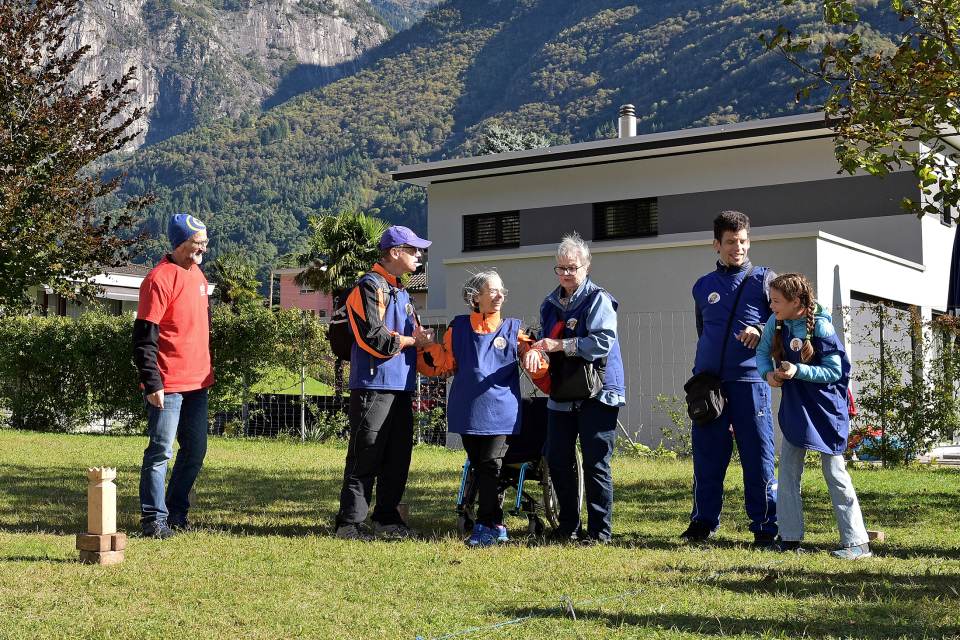 Sechs Personen stehen draußen auf einem grasbewachsenen Rasen mit Bergen im Hintergrund. Vier tragen blaue Uniformen und zwei tragen rote Hemden. Auf der Wiese sind Holzklötze angeordnet, und die Gruppe scheint sich zu unterhalten oder einer Tätigkeit nachzugehen.