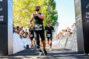Ein männlicher Athlet mit einer Beinprothese läuft bei einem Triathlon auf die Ziellinie zu, gefolgt von anderen Teilnehmern, während Zuschauer an einem sonnigen Tag hinter Absperrungen jubeln.