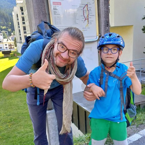 Ein lächelnder Mann mit Brille und Schal zeigt den Daumen nach oben, daneben ein Junge mit blauem Helm und Brille. Sie stehen im Freien auf einer Wiese in der Nähe eines Gebäudes und schauen in die Kamera.