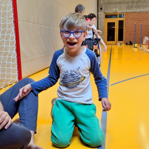 Ein lächelnder Junge mit einer blauen Brille und einem Planetenhemd kniet auf einem leuchtend gelben Turnhallenboden, während im Hintergrund andere Menschen und Turngeräte zu sehen sind.
