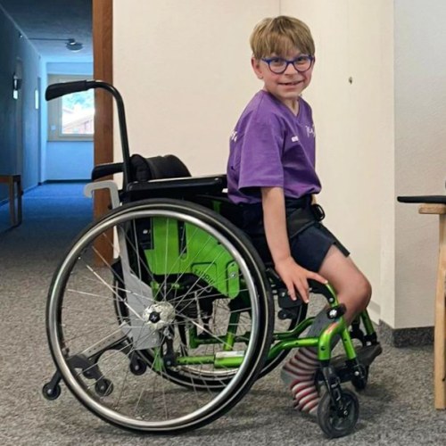 Ein Junge mit Brille, lila Hemd und gestreiften Socken lächelt, während er in einem grünen Rollstuhl in einem Haus sitzt.