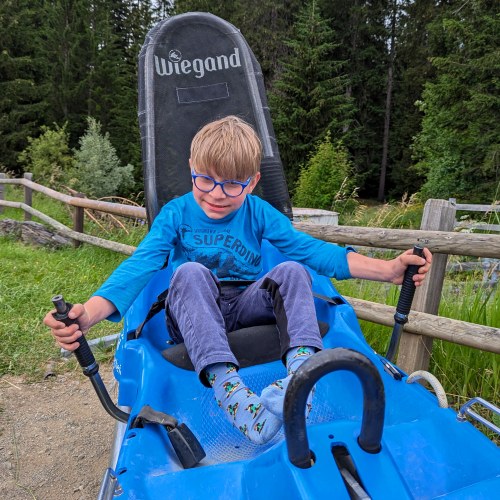 Ein kleiner Junge mit Brille sitzt in einem blauen Outdoor-Fahrzeug und hält sich an zwei Griffen fest. Er lächelt und trägt ein blaues Hemd, eine graue Hose und Socken mit Mustern. Im Hintergrund sind Bäume und ein Holzzaun zu sehen.