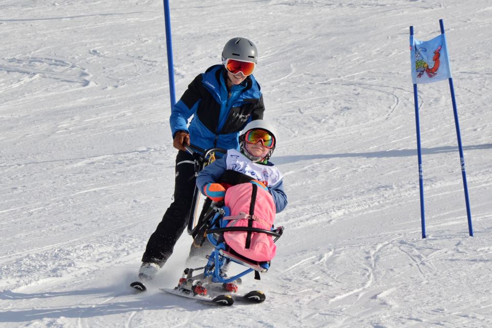 Ein Skilehrer führt ein Kind auf einem Sitz-Ski einen verschneiten Hang zwischen Fahnenmarkierungen hinunter. Das Kind ist in eine rosafarbene Decke gehüllt und beide tragen Helme und Skibrillen.