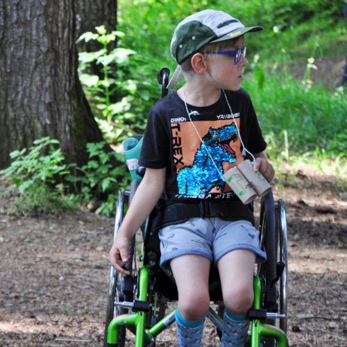 Ein kleiner Junge mit Brille, Mütze und T-Rex-Shirt sitzt in einem grünen Rollstuhl im Freien. Er hält ein gefaltetes Papier in der Hand und blickt zur Seite. Bäume und Grünzeug umgeben ihn.