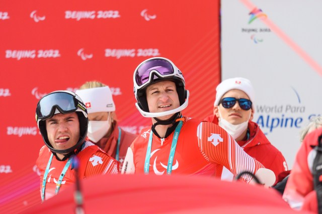 Vier Skifahrer in rot-weißen Uniformen stehen zusammen, einige tragen Helme und Skibrillen, und im Hintergrund ist das rote Peking 2022 zu sehen. Eine Person im Hintergrund trägt eine Maske.