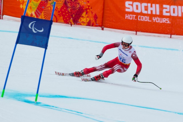 Ein rot-weiß gekleideter paralympischer Alpinskifahrer fährt bei den Winter-Paralympics in Sotschi 2014 um ein Tor herum, während im Hintergrund das orangefarbene Banner der Veranstaltung zu sehen ist.