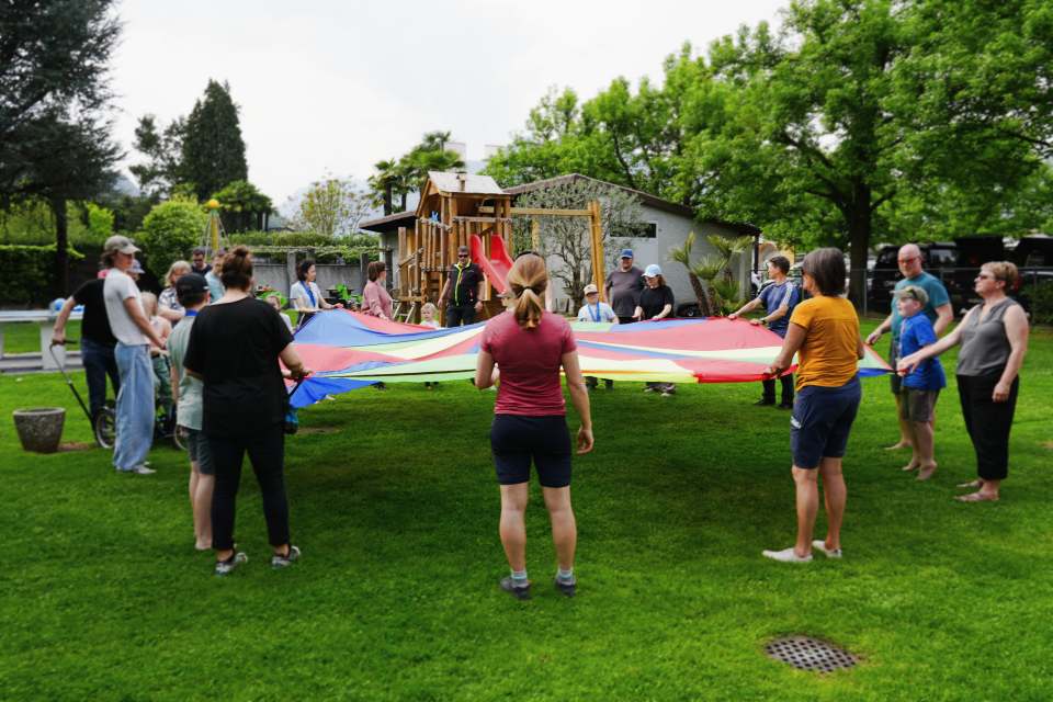 Eine Gruppe von Menschen steht in einem grasbewachsenen Hof und hält die Ränder eines großen, bunten Fallschirms, der in einem Kreis ausgebreitet ist. Im Hintergrund sind Spielgeräte und Bäume zu sehen, und das Wetter scheint sonnig und angenehm zu sein.