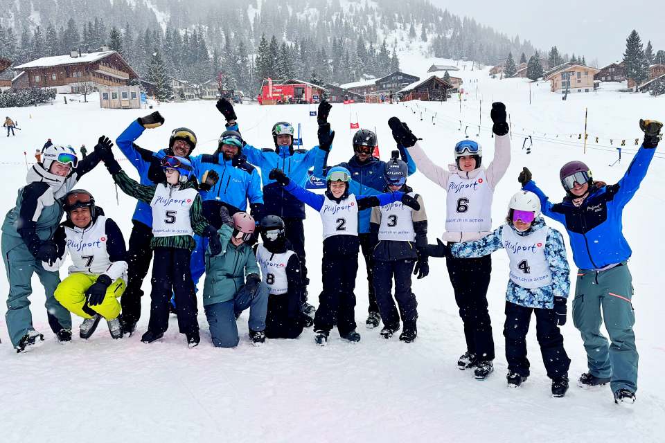 Eine Gruppe von Skifahrern, die Helme, Winterausrüstung und nummerierte Westen tragen, posieren gemeinsam auf einer verschneiten Piste, heben ihre Arme und lächeln. Im Hintergrund sind schneebedeckte Bäume und Skihütten zu sehen.