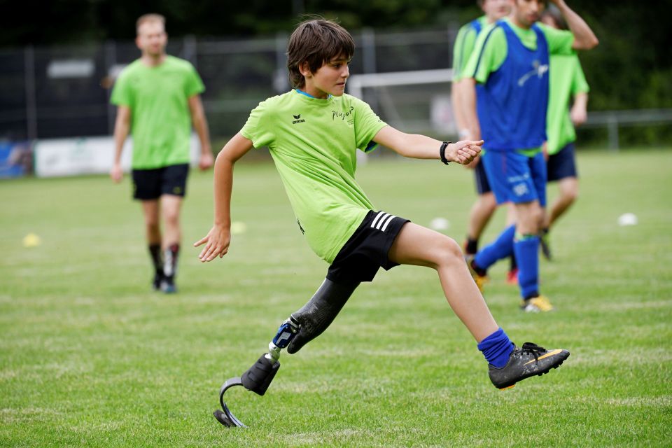 Ein Junge mit einer Beinprothese spielt Fußball auf einer Wiese, er trägt ein grünes Hemd und blaue Socken. Andere Spieler in ähnlichen Uniformen sind im Hintergrund zu sehen.