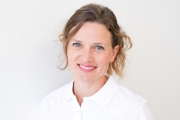 Eine Frau mit gewelltem hellbraunem Haar, die ein weißes Hemd mit Kragen trägt, lächelt in die Kamera vor einem schlichten weißen Hintergrund.