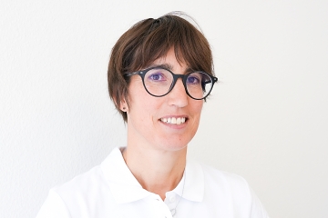 Eine Person mit kurzen braunen Haaren und Brille lächelt in die Kamera. Sie trägt ein weißes Hemd mit Kragen und steht vor einem schlichten weißen Hintergrund.