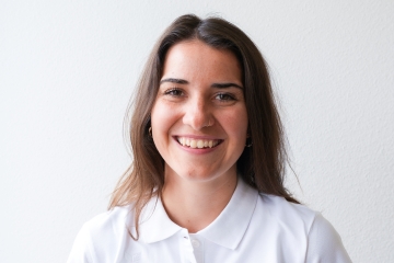 Eine junge Frau mit langen braunen Haaren und einem weißen Hemd mit Kragen lächelt vor einem schlichten weißen Hintergrund herzlich in die Kamera.
