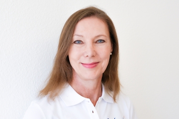Eine Frau mit glattem, hellbraunem Haar und blauen Augen lächelt in die Kamera. Sie trägt ein weißes Hemd mit Kragen und steht vor einem schlichten weißen Hintergrund.