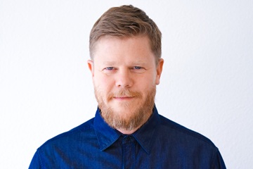 Ein Mann mit kurzen hellbraunen Haaren und Bart, der ein dunkelblaues Hemd mit Knöpfen trägt, steht vor einem schlichten weißen Hintergrund und blickt mit ausdruckslosem Gesicht direkt in die Kamera.