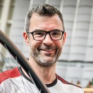 Ein Mann mittleren Alters mit kurzen dunklen Haaren, einer Brille und einem Bart lächelt in die Kamera. Er trägt ein weißes und rotes Hemd, und im Vordergrund ist ein Teil eines Fahrrads zu sehen. Der Hintergrund scheint ein Innenraum zu sein.