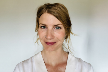 Eine Frau mit hellbraunem Haar und heller Haut lächelt in die Kamera. Sie trägt ein weißes Oberteil und einen Nasenring. Der Hintergrund ist schlicht und leicht verschwommen.