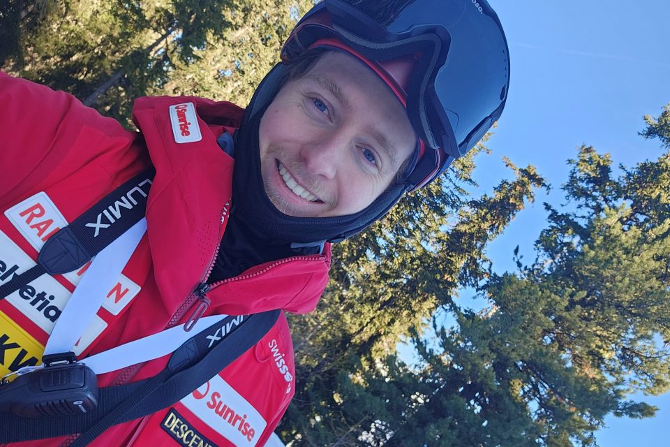 Eine Person in einer roten Skijacke und einem schwarzen Skihelm lächelt im Freien, im Hintergrund sind Bäume und ein strahlend blauer Himmel zu sehen.