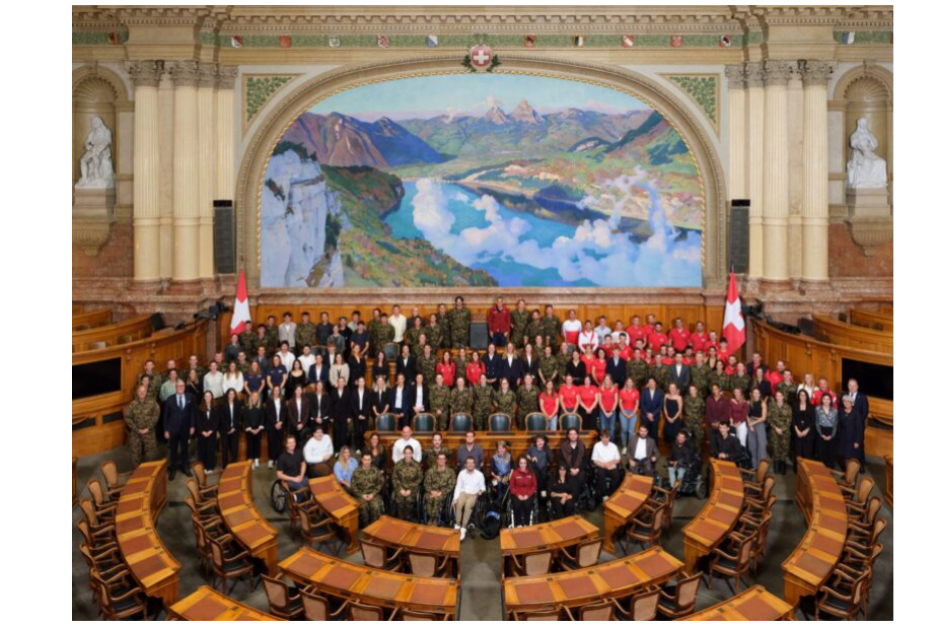 Eine große Gruppe von Menschen posiert für ein Foto im halbrunden Saal des Schweizer Nationalrats, an dessen Rückwand sich ein großes Wandgemälde mit einer Berglandschaft befindet.