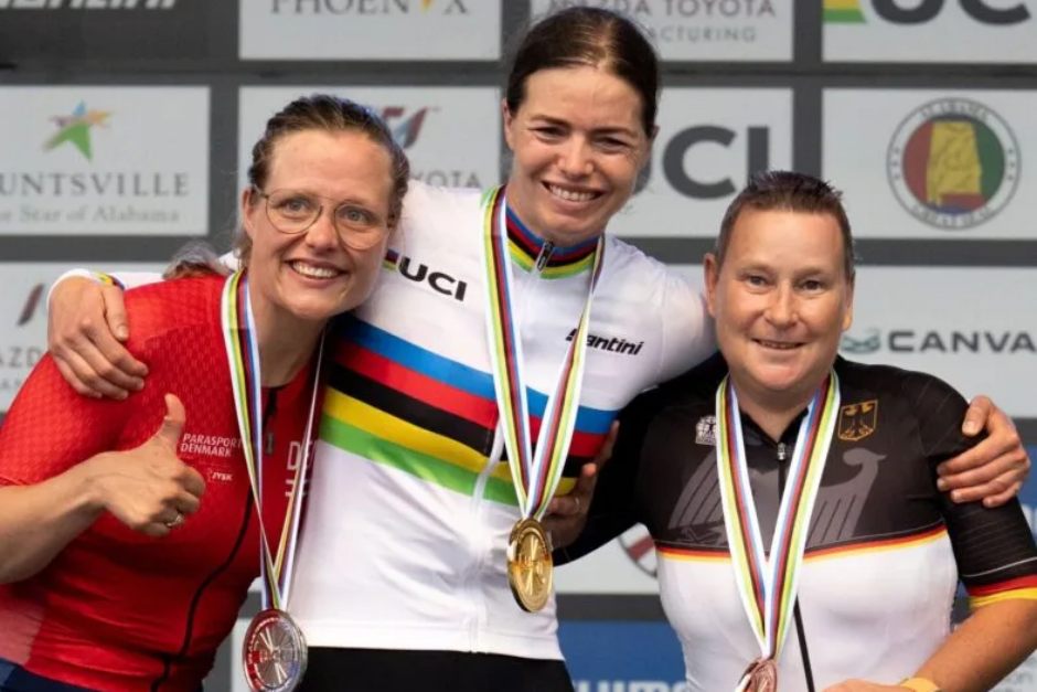 Drei Radfahrerinnen stehen lächelnd und mit Medaillen behängt auf einem Podium. Die Athletin in der Mitte trägt ein Regenbogentrikot und eine Goldmedaille, flankiert von der Silber- und der Bronzemedaillengewinnerin. Im Hintergrund sind Sponsorenlogos zu sehen.