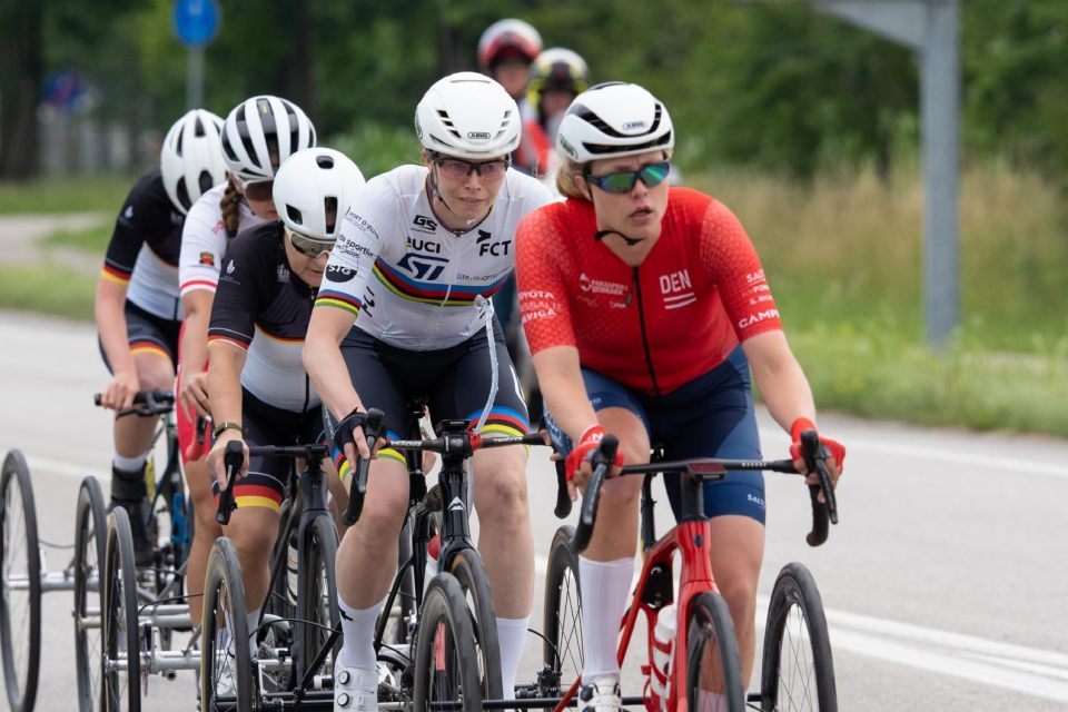 Eine Gruppe von Radfahrern, die Helme und Rennbekleidung tragen, fährt in einer Reihe auf einer Straße, wobei ein Radfahrer im roten Trikot vorne fährt und die anderen dicht hinter ihm herfahren.