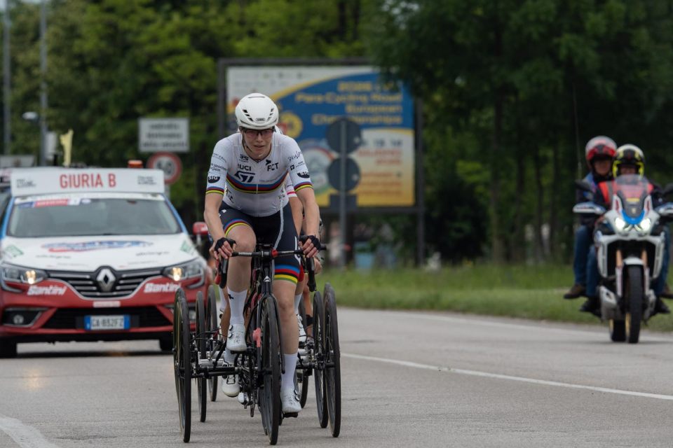 Ein Radfahrer mit weißem Helm und Rennbekleidung fährt auf einer Straße ein dreirädriges Handbike, gefolgt von einem Begleitwagen und einem Motorrad; im Hintergrund sind Bäume und Schilder zu sehen.