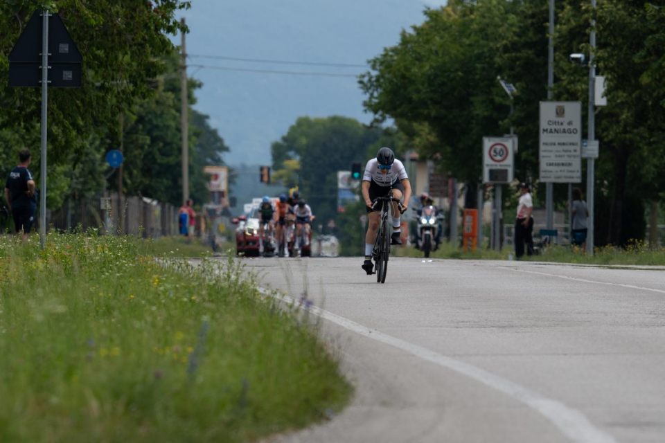 Ein Radfahrer fährt während eines Rennens allein auf einer Straße, in der Ferne sind Begleitfahrzeuge und andere Radfahrer zu sehen. Bäume und Schilder säumen die Straße, und eine Person steht auf dem Gehweg neben einem Ortsschild mit der Aufschrift „MANIAGO“.