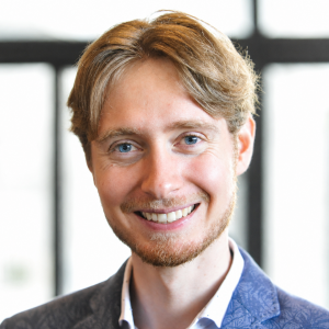 Ein junger Mann mit hellbraunen Haaren, einem kurzen Bart und blauen Augen lächelt in die Kamera. Er trägt einen blau gemusterten Blazer und ein weißes Hemd. Der Hintergrund ist leicht unscharf und zeigt große Fensterscheiben.
