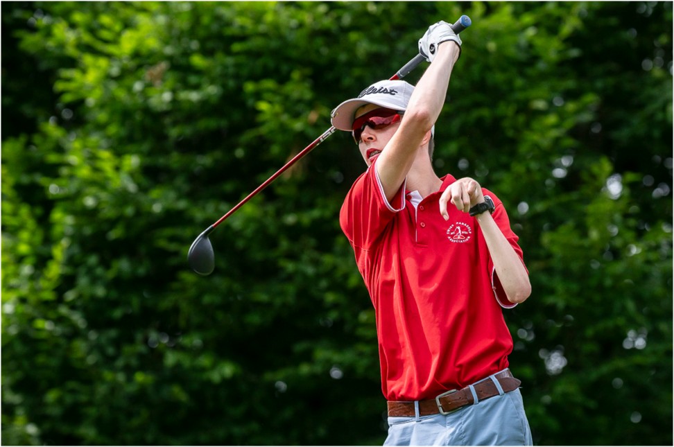 Ein Golfer mit rotem Hemd und Sonnenbrille vollführt einen Schwung mit einem Driver-Schläger, während er im Freien vor grünen Bäumen steht.