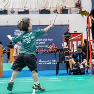 Ein Kind in einem grünen Schweiz-Trikot spielt Badminton in einer Halle und schlägt mit dem Schläger nach dem Federball. Im Hintergrund sind Funktionäre und Medienvertreter zu sehen.