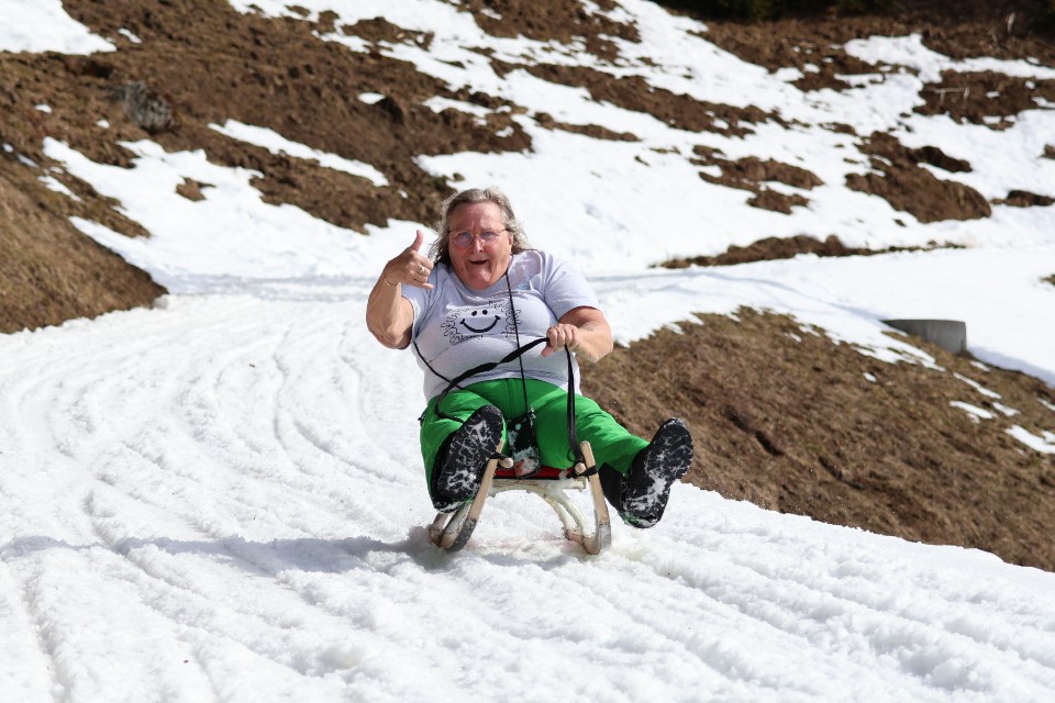 Eine ältere Frau in grüner Hose und weißem Hemd lächelt und zeigt den Daumen nach oben, während sie mit einem Schlitten einen verschneiten Hügel hinunterfährt, auf dem einige Stellen frei liegen.