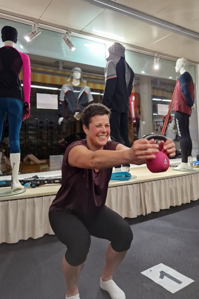 Eine Frau in Trainingskleidung geht in die Hocke, während sie eine rosafarbene Kettlebell vor sich hält, und lächelt in einem hell erleuchteten Geschäft, in dem hinter ihr Schaufensterpuppen in Sportkleidung stehen.